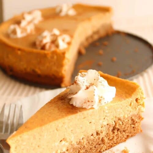 A slice of pumpkin cheesecake topped with whipped cream and a sprinkle of cinnamon is placed on a white plate with a fork. A partially cut cheesecake is in the background.