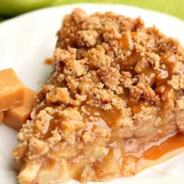 A slice of caramel apple crumble pie on a white plate, topped with caramel sauce and accompanied by caramel candy pieces.