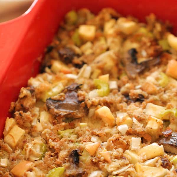 A red baking dish containing a homemade stuffing with visible chunks of bread, mushrooms, celery, and onions.