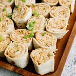 A wooden tray filled with pinwheel sandwiches made of tortillas, filled with a creamy mixture, and garnished with chopped green onions.