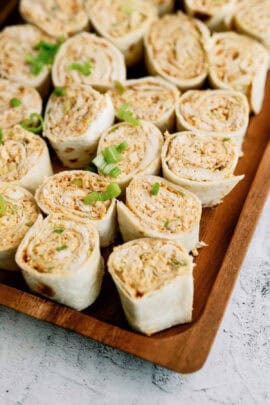 A wooden tray filled with pinwheel sandwiches made of tortillas, filled with a creamy mixture, and garnished with chopped green onions.