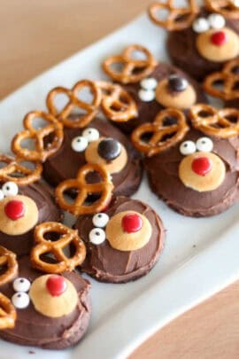 A plate of cookies decorated with chocolate, pretzels, and candies to resemble reindeer faces. The cookies have red noses, white eyes, and pretzel antlers.