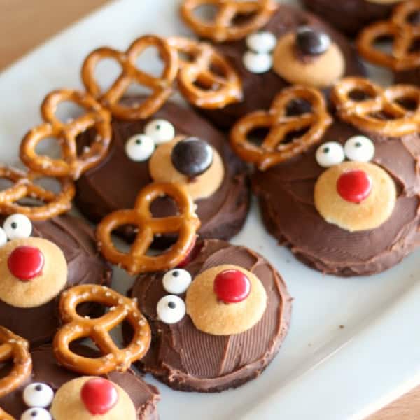 A plate of cookies decorated with chocolate, pretzels, and candies to resemble reindeer faces. The cookies have red noses, white eyes, and pretzel antlers.