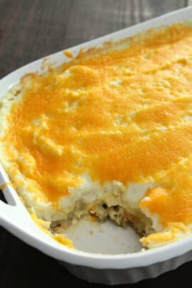 A white casserole dish filled with baked leftover thanksgiving dinner casserole topped with melted cheese, showing a portion missing.