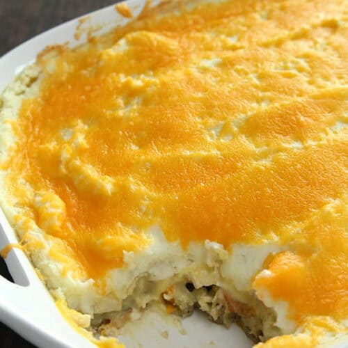 A white casserole dish filled with baked leftover thanksgiving dinner casserole topped with melted cheese, showing a portion missing.