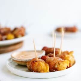 White plate with bacon-wrapped tater tots and toothpicks, accompanied by a small bowl of dipping sauce. Blurred food items in the background.