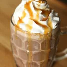 A glass mug filled with hot chocolate, topped with whipped cream, and drizzled with caramel sauce.