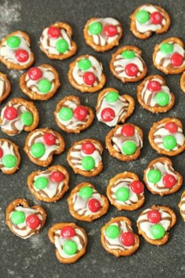 Pretzels topped with melted chocolate and red and green candies arranged on a dark surface.