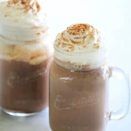 Two clear glass mugs of hot chocolate topped with whipped cream and a sprinkle of cinnamon on a light-colored surface.