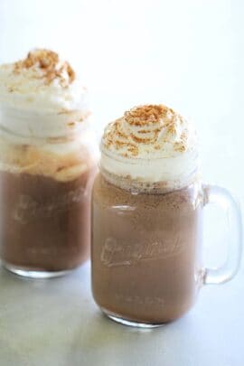 Two clear glass mugs of hot chocolate topped with whipped cream and a sprinkle of cinnamon on a light-colored surface.