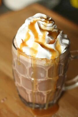 A glass mug filled with hot chocolate, topped with whipped cream, and drizzled with caramel sauce.