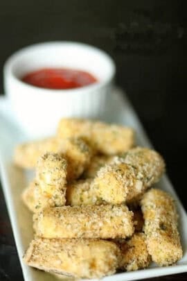 Breaded mozzarella sticks on a rectangular plate with a small white cup of red dipping sauce in the background.