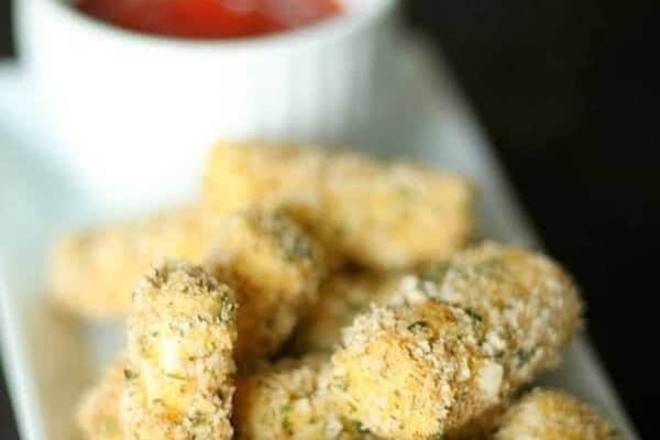 Breaded mozzarella sticks on a rectangular plate with a small white cup of red dipping sauce in the background.