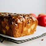 A loaf of caramel apple bread with pecans drizzled on top, placed on a cooling rack with two red apples in the background.