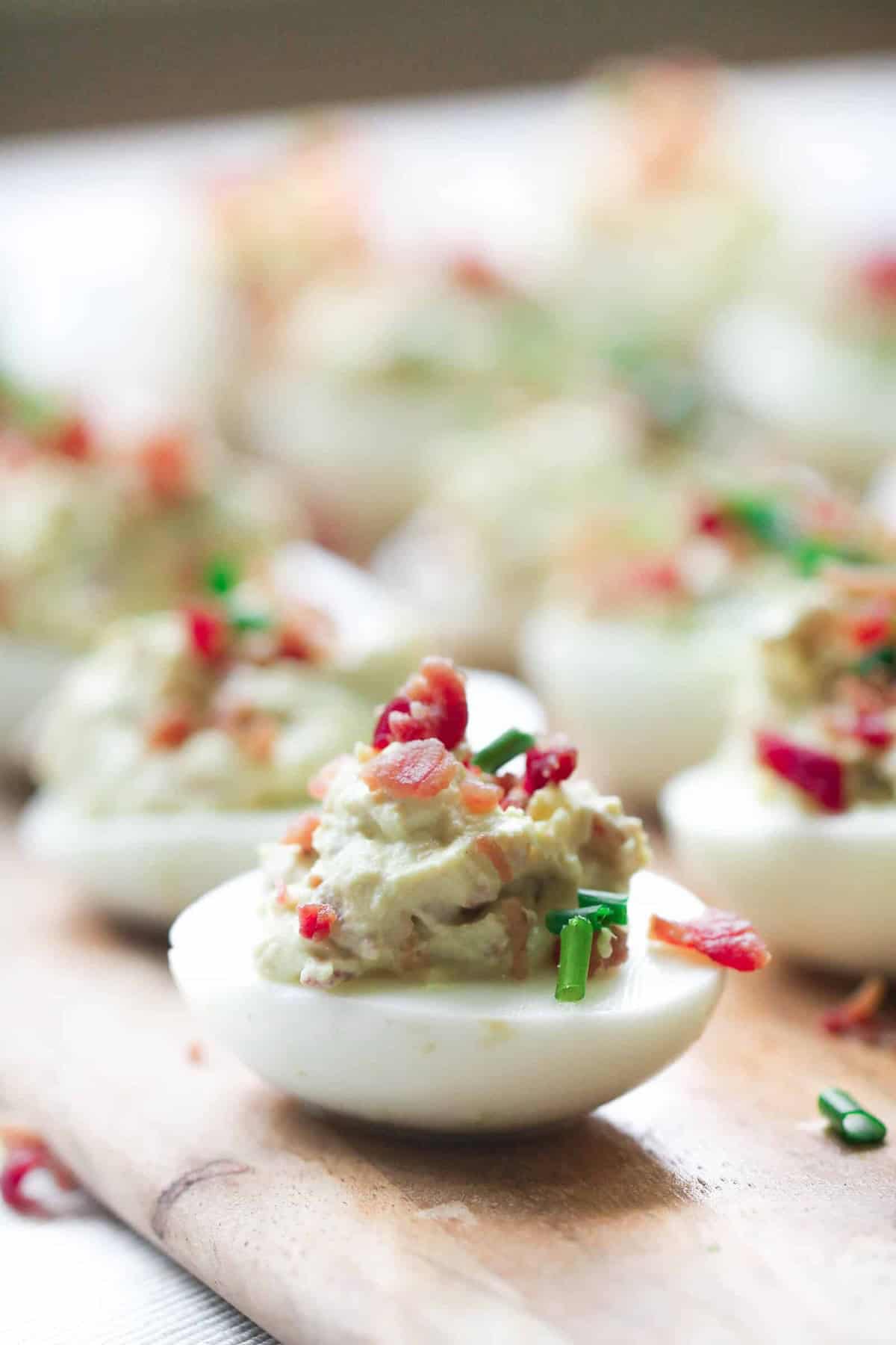 Deviled eggs topped with chopped chives and bacon are arranged on a wooden board.