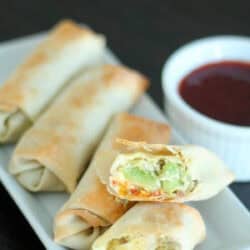 Plate with five baked avocado egg rolls, one cut open to show vegetable filling. A small dish of dipping sauce is beside them.