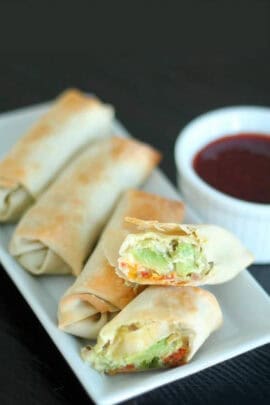 Plate with five baked avocado egg rolls, one cut open to show vegetable filling. A small dish of dipping sauce is beside them.