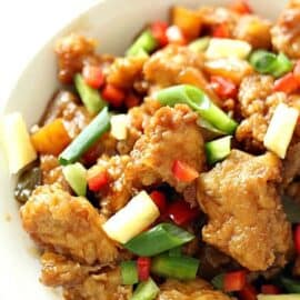 Close-up of a plate of sweet and sour chicken with colorful bell peppers, pineapple chunks, and green onions.