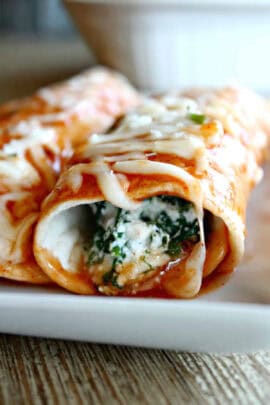 Close-up of two cheese and spinach stuffed enchiladas on a white plate, topped with tomato sauce and melted cheese.