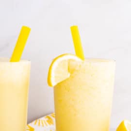 Two glasses of a yellow frozen drink with yellow straws, garnished with lemon slices.