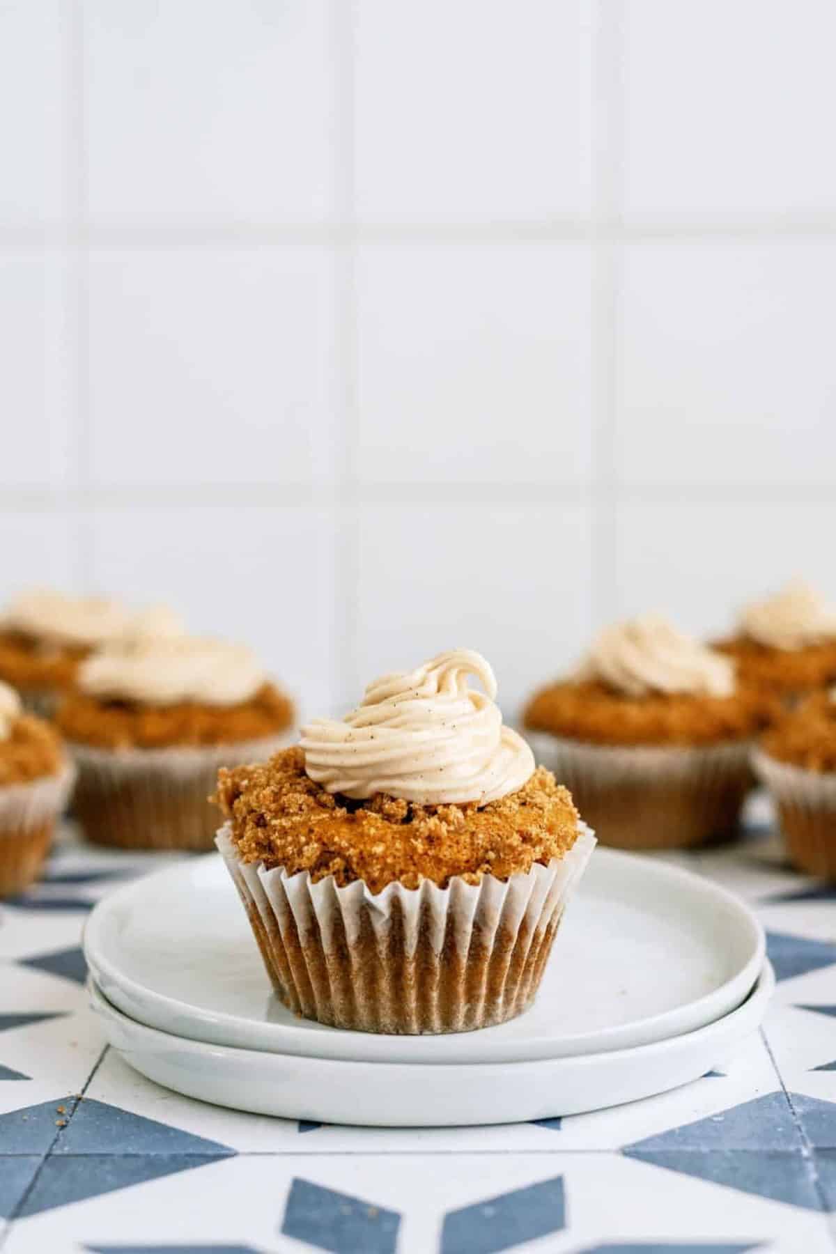 A group of six cupcakes with light brown frosting on a white plate against a tiled background.