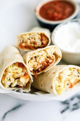 Four breakfast burritos filled with eggs, cheese, and other ingredients are displayed on a white plate. A small bowl of sour cream is next to the burritos.