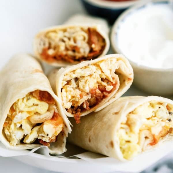 Four breakfast burritos filled with eggs, cheese, and other ingredients are displayed on a white plate. A small bowl of sour cream is next to the burritos.