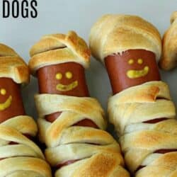 Three hot dogs wrapped in pastry dough are designed to look like mummies. Each has a smiling face made of mustard.