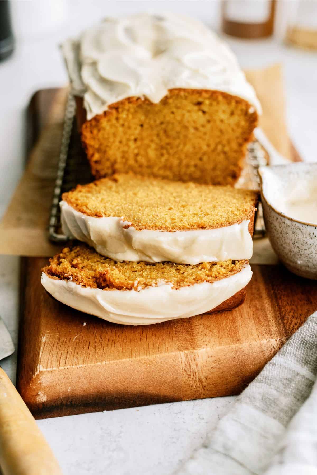 A loaf of sliced pumpkin bread with cream cheese frosting on a wooden board, next to a bowl of additional frosting.