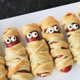 Hot dogs wrapped in dough to look like mummies, with candy eyes peeking out, arranged in a row on a white surface.