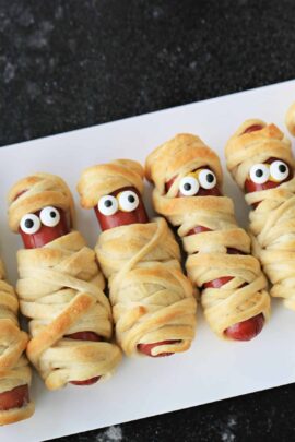 Hot dogs wrapped in dough to look like mummies, with candy eyes peeking out, arranged in a row on a white surface.