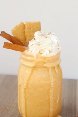 A mason jar filled with a creamy pumpkin pie milkshake, topped with whipped cream, two cinnamon sticks, and two graham crackers.