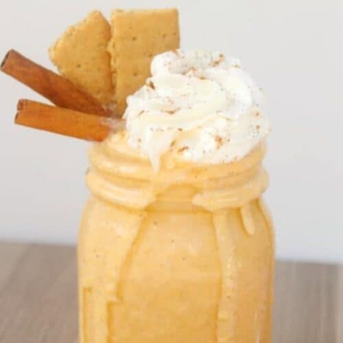 A mason jar filled with a creamy pumpkin pie milkshake, topped with whipped cream, two cinnamon sticks, and two graham crackers.