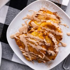 A white plate holds slices of pumpkin pull apart bread drizzled with cinnamon cream, placed on a black and white checkered tablecloth.