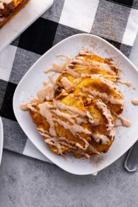 A white plate holds slices of pumpkin pull apart bread drizzled with cinnamon cream, placed on a black and white checkered tablecloth.