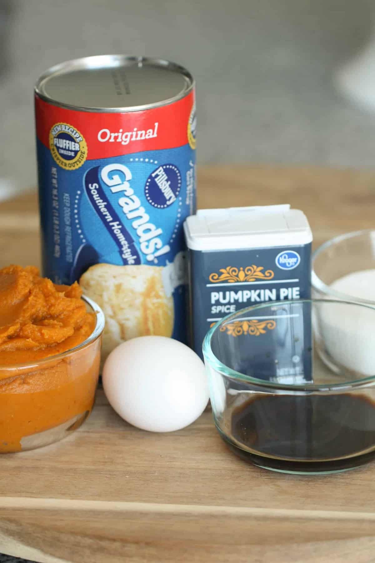 A can of biscuits, a can of pumpkin puree, an egg, a bowl of vanilla extract, a bowl of sugar, and a container of pumpkin pie spice on a wooden surface.