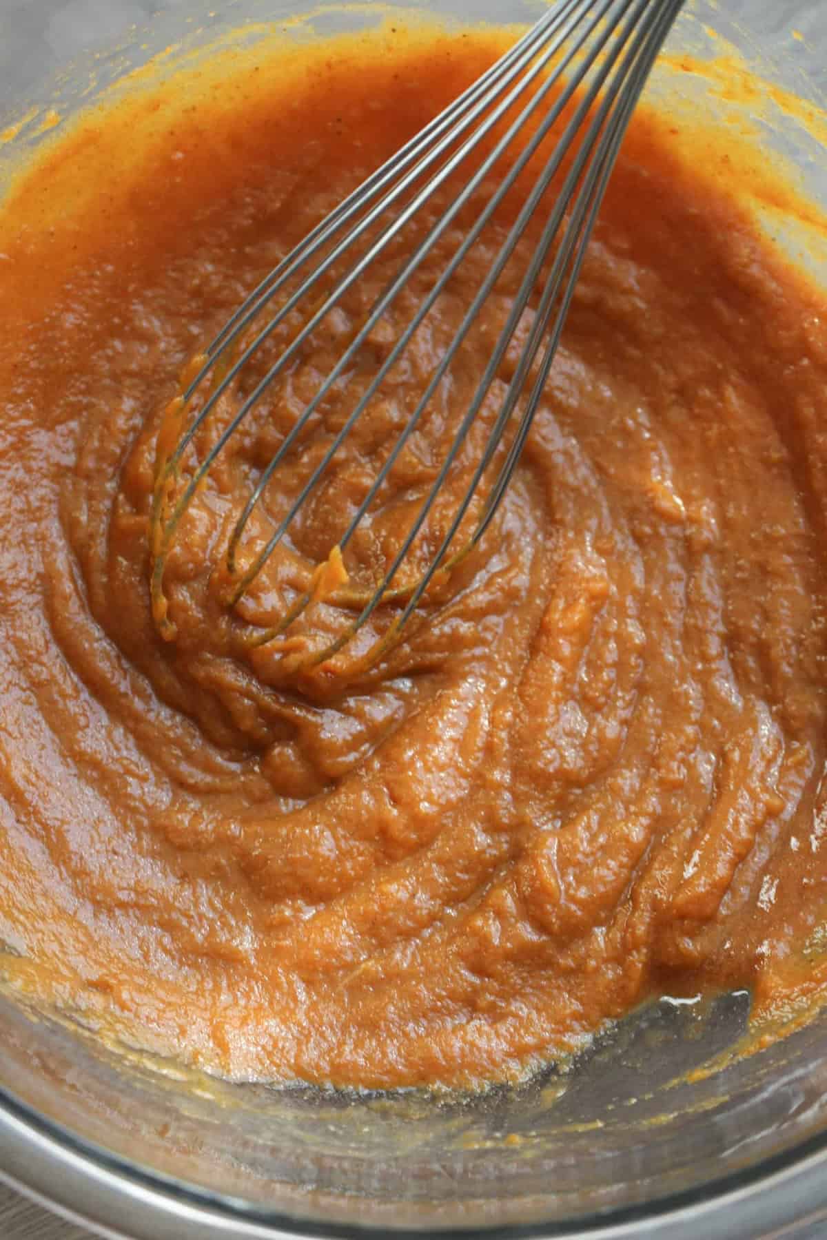 A whisk is shown in a glass bowl containing a thick orange mixture, likely a batter or purée.