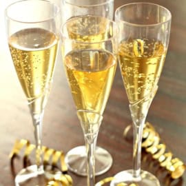 Four champagne flutes filled with a non-alcoholic holiday bubble drink are placed on a wooden table, surrounded by gold ribbon decorations.