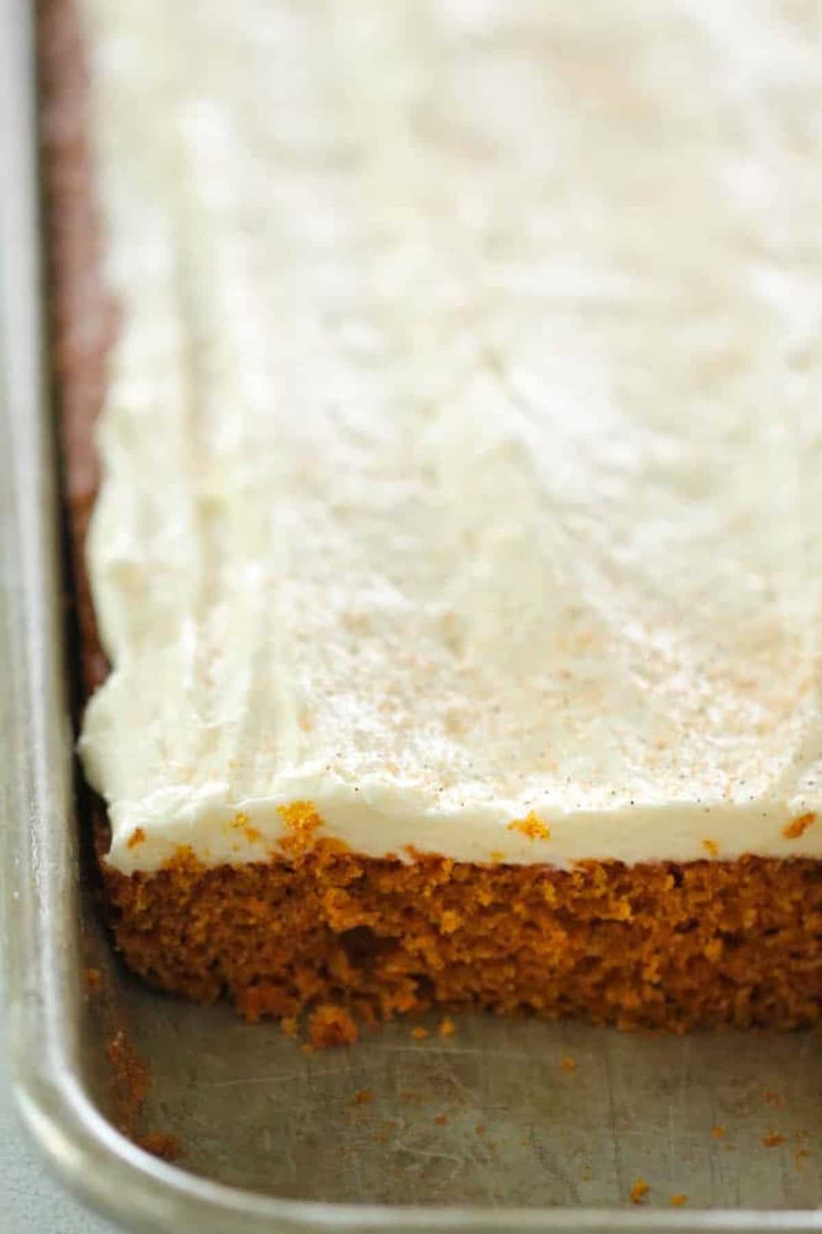 A frosted sheet cake with a cut corner revealing a moist, orange-brown interior on a metal baking tray.