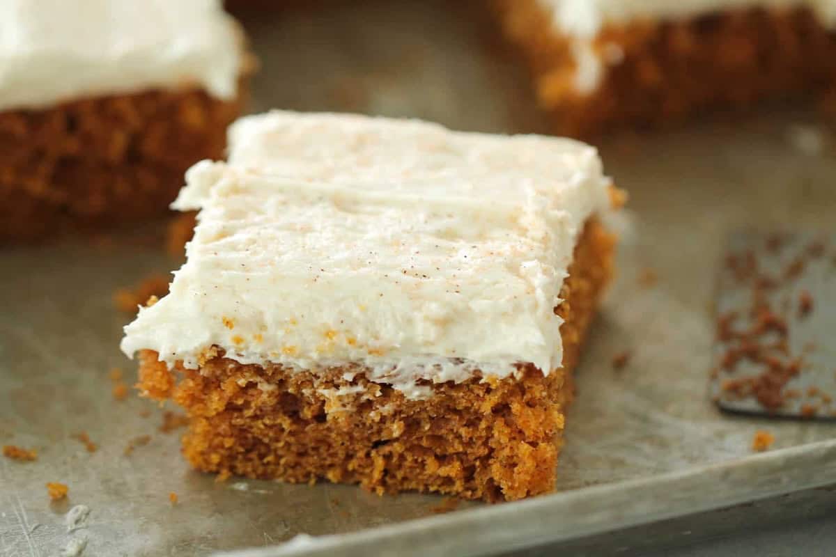 A square piece of frosted pumpkin cake with cream cheese icing on a metal baking tray, partially sliced with a spatula visible nearby.