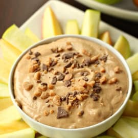 A bowl of creamy dip garnished with crushed nuts and chocolate chips is surrounded by sliced apple wedges.