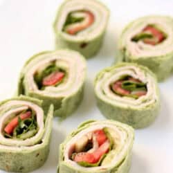 Six pieces of green tortilla wrap pinwheels filled with turkey, lettuce, tomato, and cheese are arranged on a white plate.