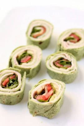 Six pieces of green tortilla wrap pinwheels filled with turkey, lettuce, tomato, and cheese are arranged on a white plate.