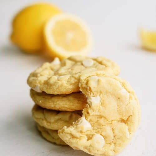 Lemon Cheesecake Pudding Cookies stacked with on cookie leaning on the stack missing a bite. Fresh lemons in the background.