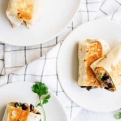 Three grilled burritos on white plates with a sprig of cilantro, placed on a checkered cloth background.