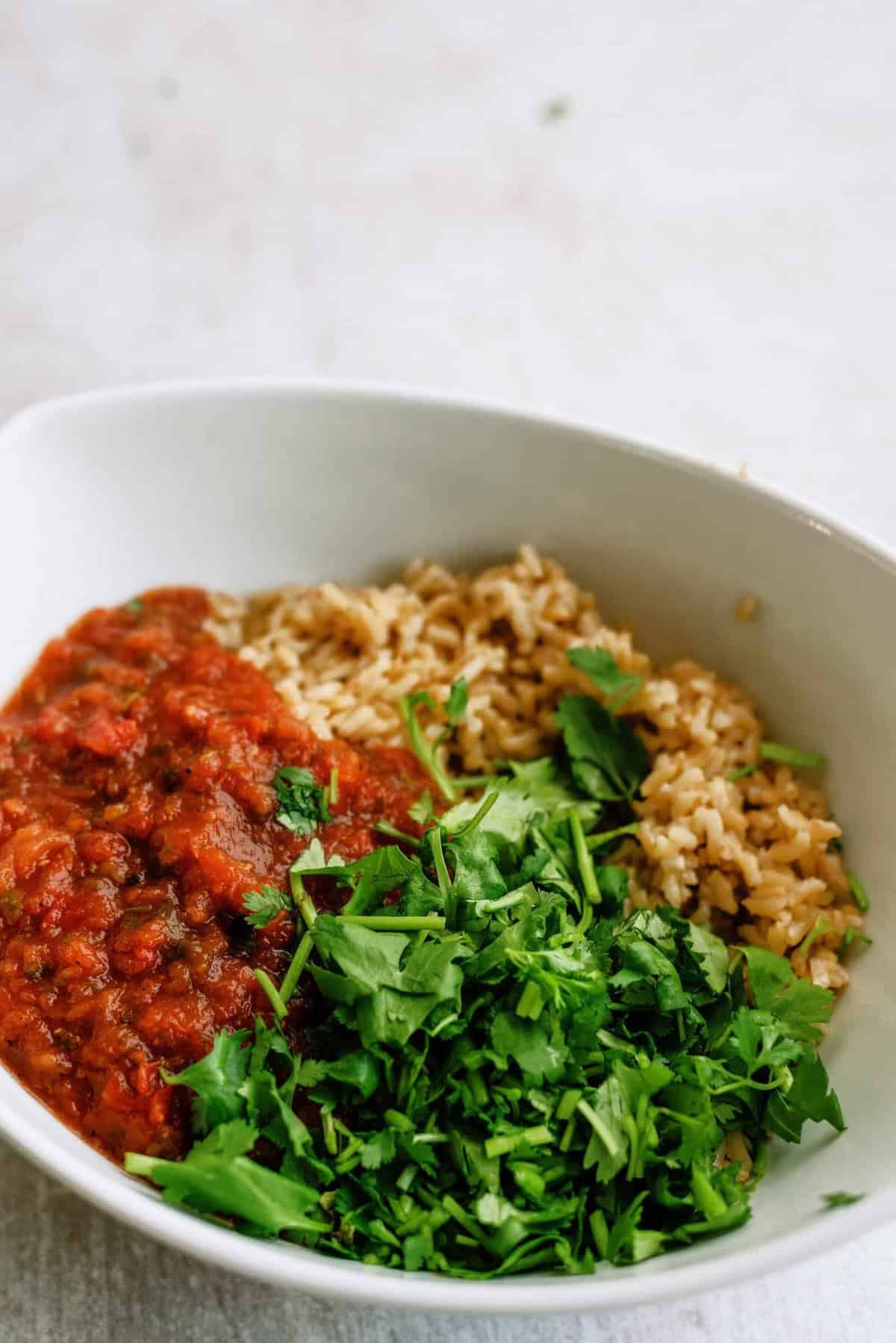 A bowl containing cooked brown rice, red tomato sauce, and fresh chopped green herbs.