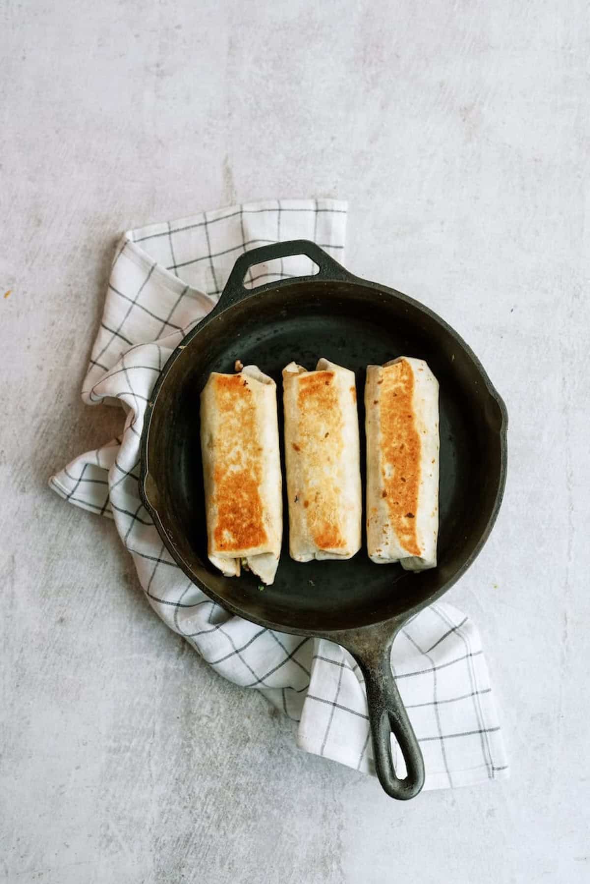 Three burritos are neatly arranged in a black skillet, set on a white and black checkered cloth, against a light gray background.