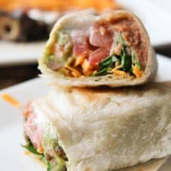 Close-up of a wrap cut in half, showing avocado, lettuce, and shredded cheese filling. Placed on a white plate with a blurred background.