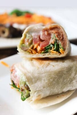 Close-up of a wrap cut in half, showing avocado, lettuce, and shredded cheese filling. Placed on a white plate with a blurred background.