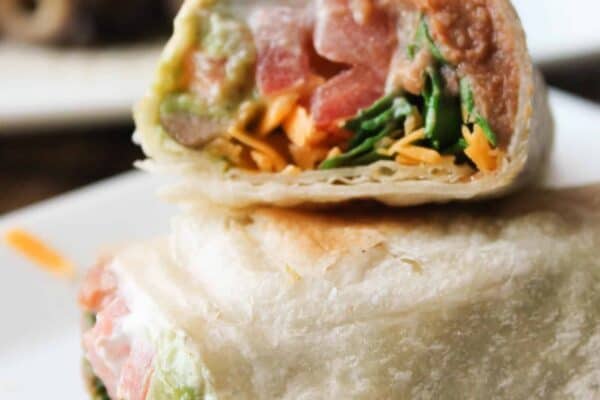 Close-up of a wrap cut in half, showing avocado, lettuce, and shredded cheese filling. Placed on a white plate with a blurred background.